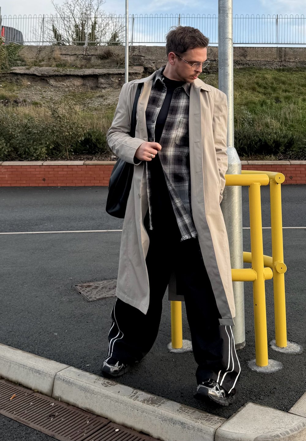 Trench-coat beige porté sur une chemise noire et une flanelle à carreaux. Pantalon large noir avec des bandes blanches sur les côtés, et baskets grises. Sac à bandoulière noir.