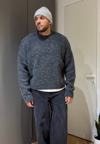 Homme portant un bonnet en maille gris, un pull duveteux gris foncé et un pantalon ample foncé, debout à l'intérieur près d'une porte fermée et d'une plante.