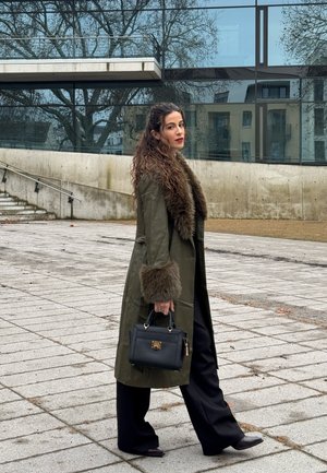 Mujer con cabello largo y rizado, vestida con un abrigo verde con ribete de piel y pantalones negros, sosteniendo un bolso negro, caminando al aire libre cerca de un edificio moderno.