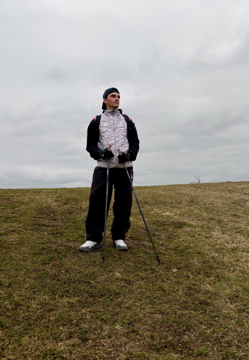 Un homme en tenue de randonnée avec des bâtons de trekking se tient sur une colline herbeuse sous un ciel nuageux, regardant sur le côté.