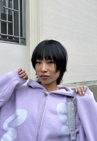 Jeune personne aux cheveux courts noirs portant un sweat à capuche lavande, montrant une manucure détaillée rose et or, debout devant un mur clair et une grille de fenêtre en métal.