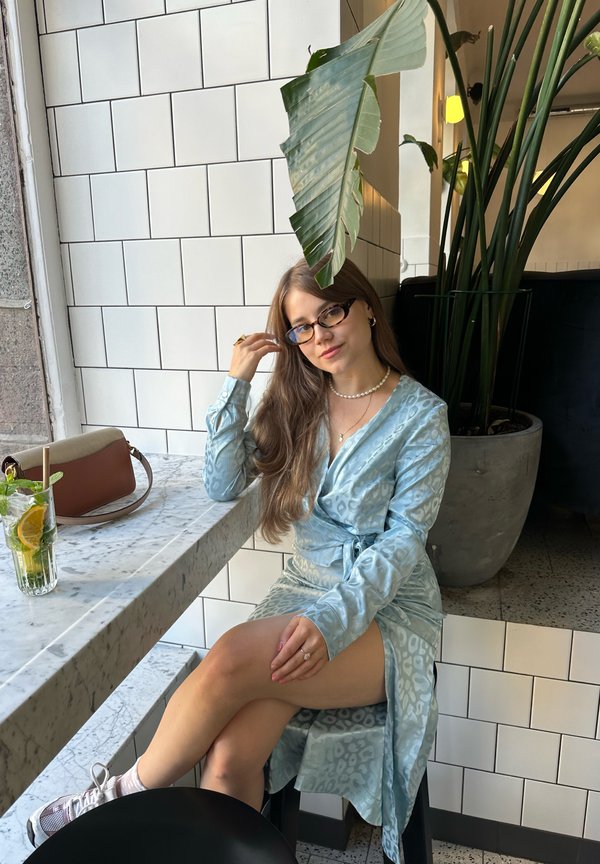 Jasnoniebieska sukienka w wzory z długimi rękawami, ściągnięta w talii. Gładki materiał, w połączeniu z koktajlem i brązową torebką na marmurowym stole.