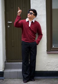 Polo roșu închis cu mânecă lungă și guler alb, asortat cu pantaloni negri. Subiectul poartă ochelari de soare negri, cu rame pătrate, stând lângă o ușă verde.