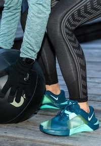 Türkizkék és zöld sportcipők texturált felsőrésszel, fekete mintás leggingsszel és fekete medicinlabdával párosítva. Egy kézen kesztyűt viselnek.