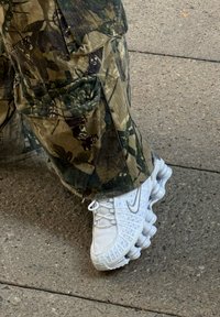 Adidași Nike albi cu un superior din plasă texturată și talpă cu amortizare vizibilă, asortați cu pantaloni cargo de camuflaj, dotati cu multiple buzunare.