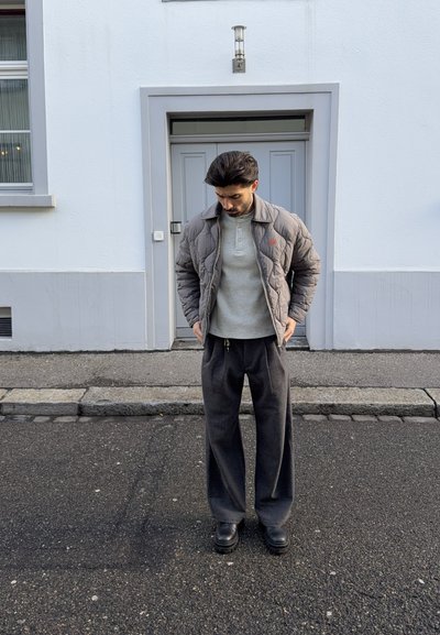 Hombre con chaqueta acolchada gris, camisa gris claro, pantalones anchos gris oscuro y zapatos negros, de pie sobre asfalto frente a una puerta cerrada.