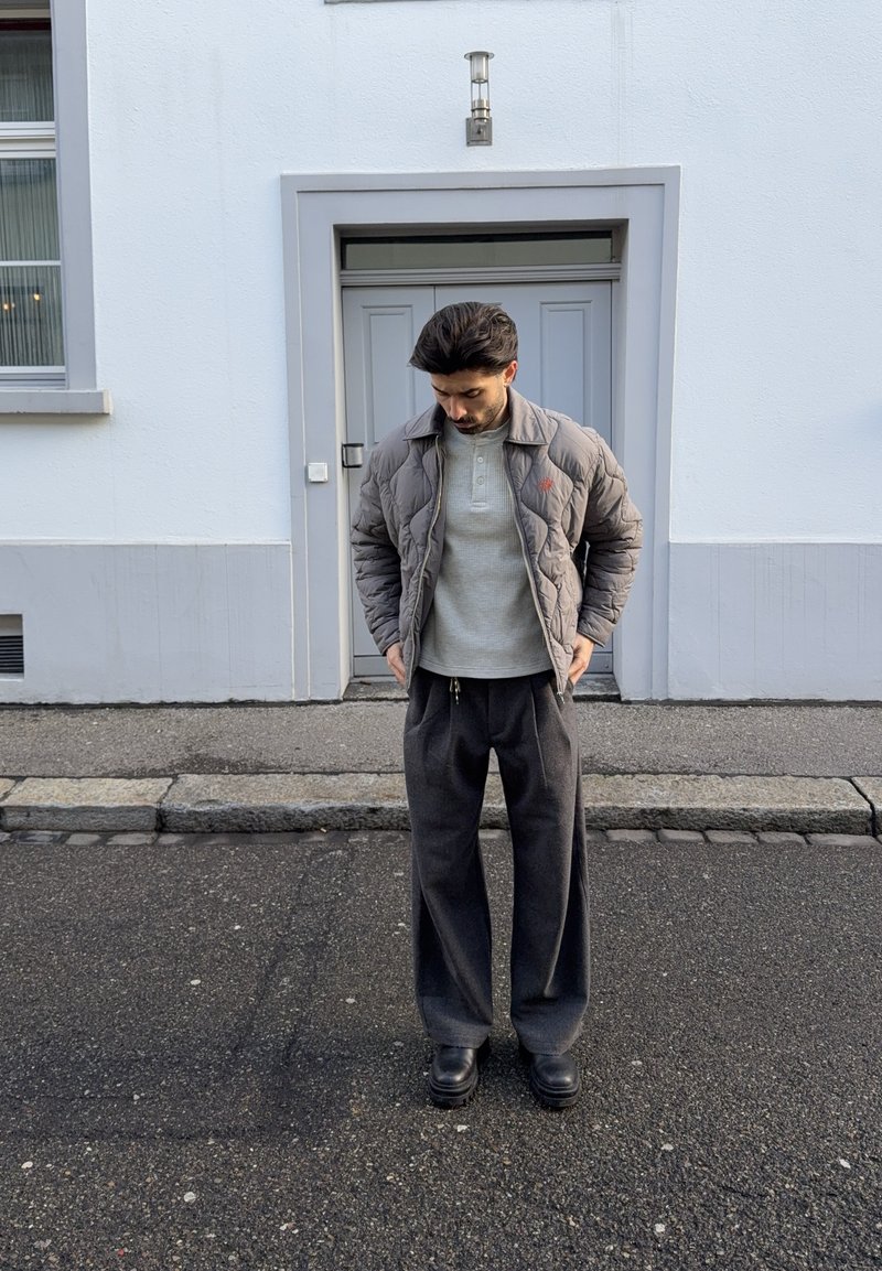 Muž v šedej prešívanej bunde, svetlosivej košeli, tmavosivých širokých nohaviciach a čiernych topánkach stojaci na asfalte pred zatvorenými dverami.