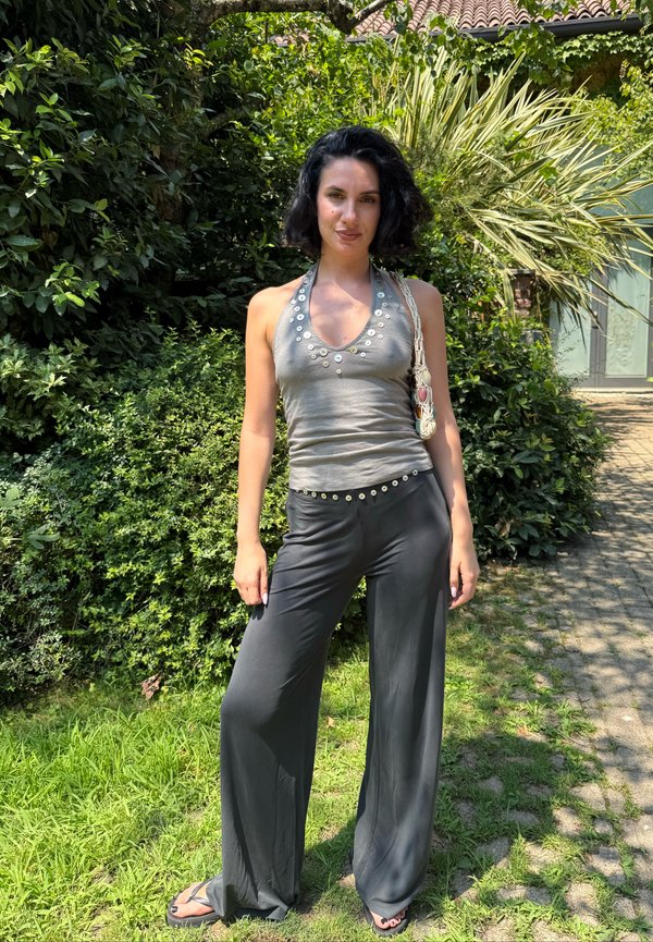 Femme aux cheveux courts et foncés portant un haut gris à col halter et un pantalon large noir, debout sur l'herbe dans un jardin ensoleillé avec des plantes vertes denses derrière.