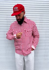 Camisa de manga longa xadrez vermelho e branco com dois bolsos frontais e detalhes brancos nas mangas, combinada com um boné vermelho com um logótipo.