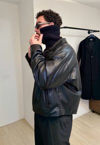 Homme portant une veste en cuir noire et un épais col roulé noir, ajustant son col à l'intérieur, un manteau sombre accroché à un porte-manteau mural derrière lui.