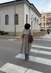 Rutig trenchcoat med bälte vid midjan, bärs över blå utsvängda jeans. Mörkbrun axelväska, som korsar en randig övergångsställe.