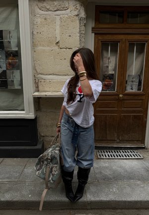 Mujer de pie en la acera con ropa informal sosteniendo una bolsa de lona con estampado, con fondo de pared de piedra y puerta de madera.