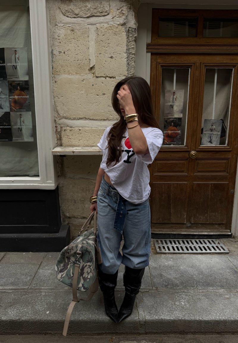 Woman standing on sidewalk in casual outfit holding a patterned duffel bag, with stone wall and wooden door background.