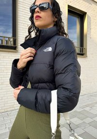 Μαύρο μπουφάν με ψηλό γιακά και λογότυπο, συνδυασμένο με ανάγλυφο ελαιοπράσινο σύνολο και λευκή τσάντα ώμου.