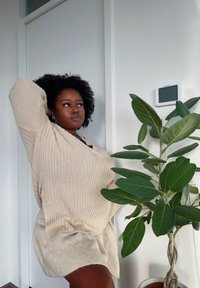 A woman with curly hair wearing a beige sweater and skirt poses next to a green potted plant in a bright, minimalist room.