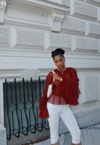 Rote plissierte Bluse mit weit ausgestellten Ärmeln, weiße schmal geschnittene Hose und eine kleine handtasche in Creme. Texturierte Wand im Hintergrund mit schwarzem Geländer.