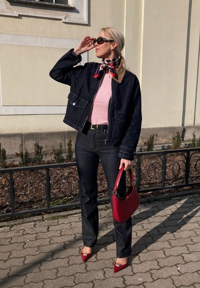 Chaqueta de denim con un collar marrón, top de canalé rosa, jeans azul oscuro y tacones rojos de punta. Sostiene un bolso rojo. Cabello largo y rubio.