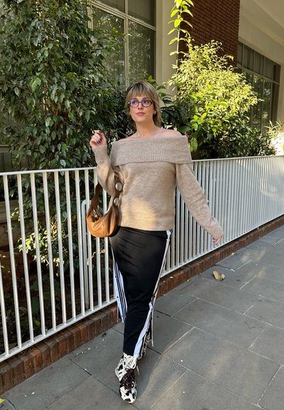 Suéter beige de un solo hombro, pantalones negros de pierna ancha con rayas blancas, bolso marrón y zapatos con patrón en blanco y negro contra un fondo de vegetación.