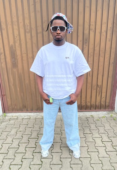 Camiseta blanca de gran tamaño con rayas horizontales y logo, jeans baggy de color azul claro, gorra de estampado, gafas de sol blancas, objeto verde en la cintura.