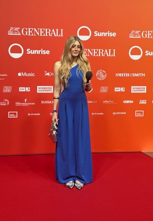 Una mujer en un mono azul cobalto sostiene un micrófono. Lleva gafas, zapatos plateados con lazos y lleva un bolso metálico contra un fondo rojo.