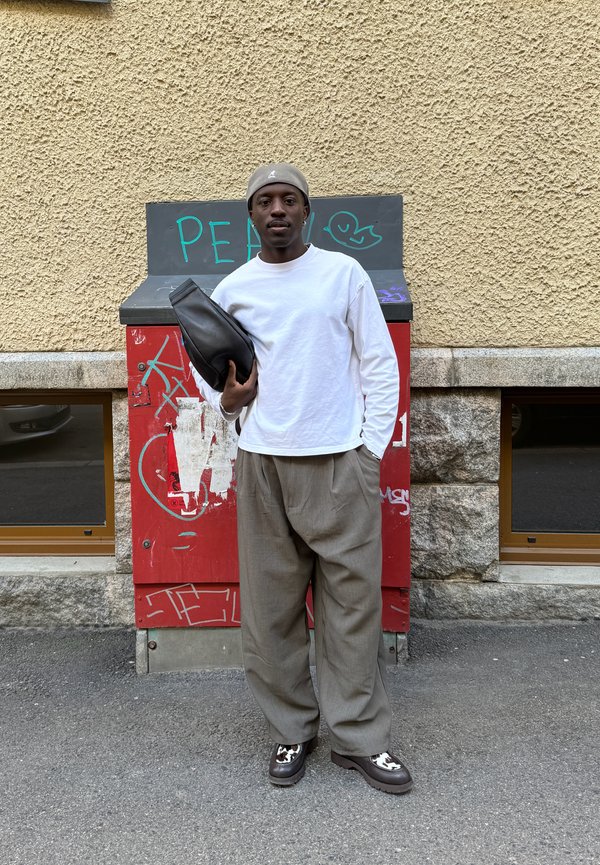 Hombre con camisa blanca de manga larga, pantalones grises y gorra beige, de pie en la calle sosteniendo una bolsa negra, con una caja cubierta de grafiti rojo detrás de él.