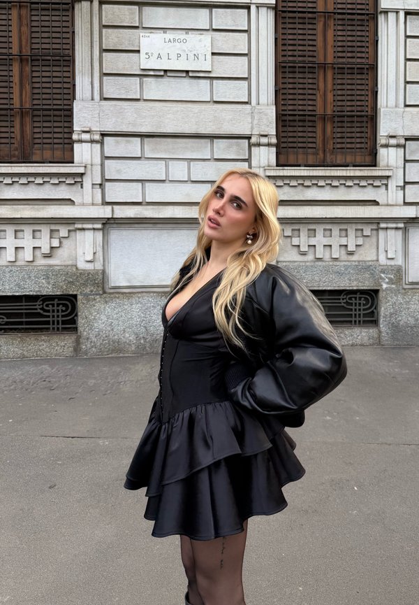 Schwarzes, geschichtetes Kleid mit tailliertem Oberteil und voluminösem Rock, kombiniert mit einer puffigen schwarzen Lederjacke. Glatte Textur und elegantes Design.