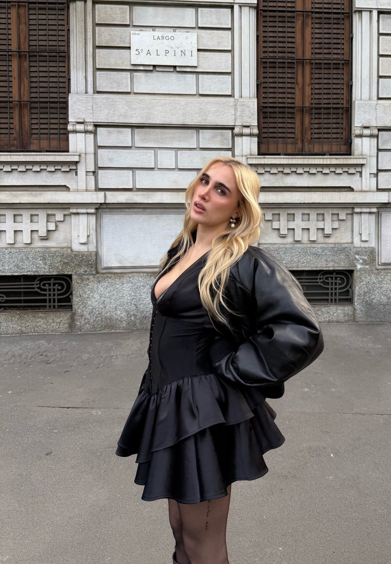 Robe noire superposée avec un corsage ajusté et une jupe à volants, associée à une veste en cuir noire bouffante. Texture lisse et design élégant.