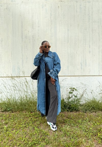 Trench de mezclilla sobre una blusa negra, pantalones de pierna ancha grises y zapatillas negras con detalles en blanco. Unas gafas de sol y un bolso negro completan el look.