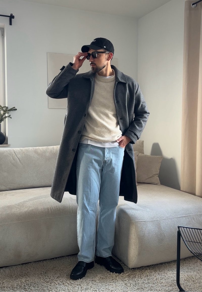 Homme portant un pardessus gris, un pull beige, un jean bleu clair, des chaussures noires, une casquette et des lunettes de soleil, debout dans un salon moderne.