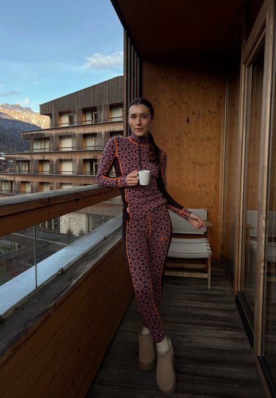 Conjunto de ropa deportiva con base burdeos y patrones geométricos, acentuado en naranja, con cuello alto y diseño ajustado. Balcón de madera visible.