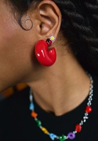 Oreille avec trois boucles d'oreilles : grand anneau rouge incurvé, petite coccinelle, et petite marguerite blanche, avec un collier de perles multicolores sur un haut noir.