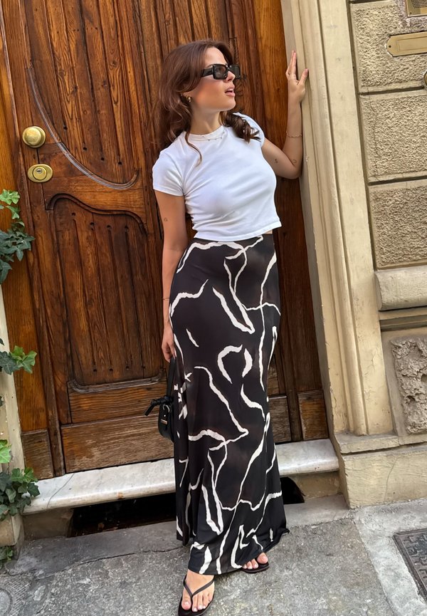 T-shirt bianca croppata a maniche corte, gonna maxi nera con motivi astratti bianchi, ciabatte nere e borsa nera piccola.
