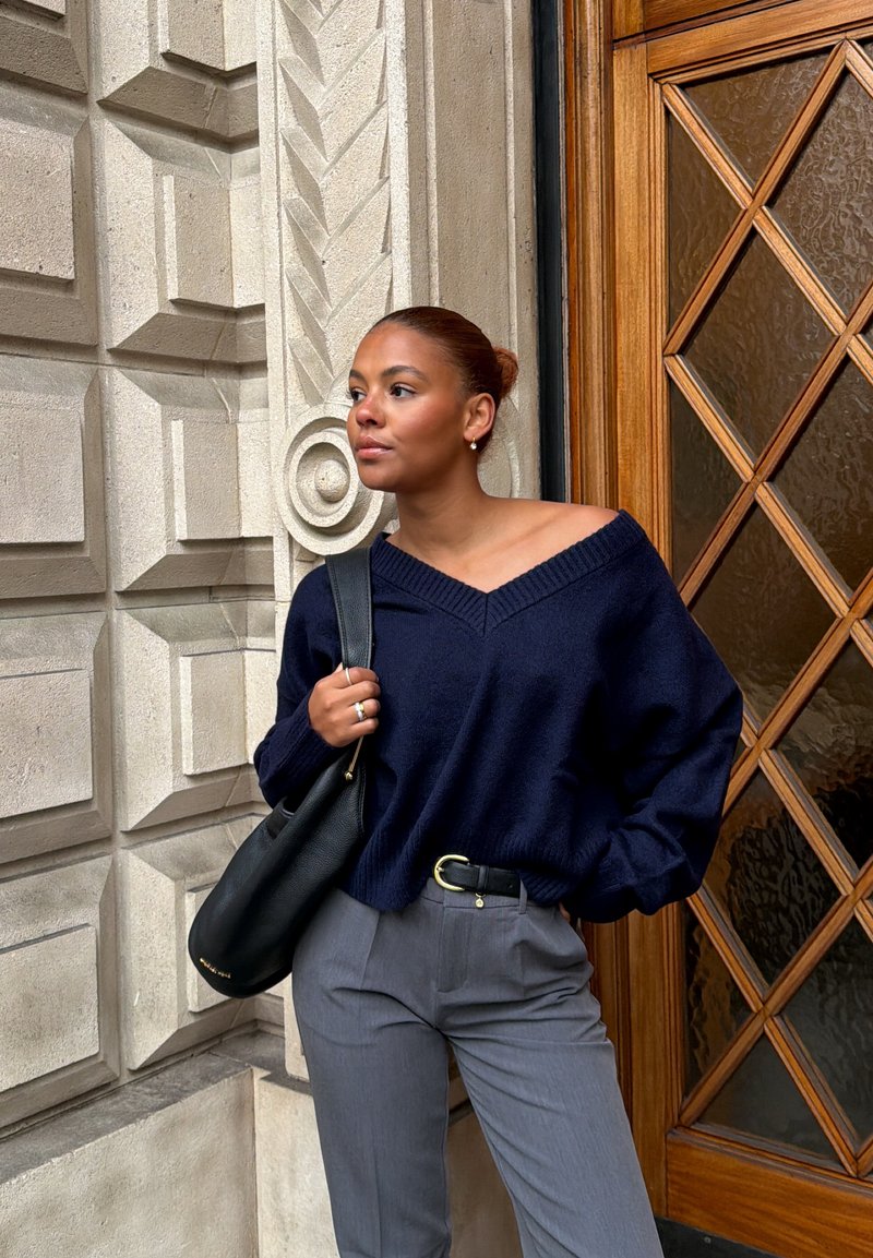 Jeune femme aux cheveux roux attachés en chignon, portant un pull marine et un pantalon gris, debout près d'un mur en pierre décoratif et d'une porte en verre avec encadrement en bois.