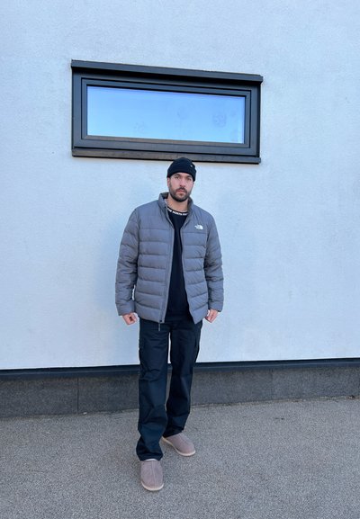 Chaqueta acolchada gris, camisa negra, pantalones cargo y pantuflas marrón claro. De pie contra una pared blanca con una ventana rectangular.