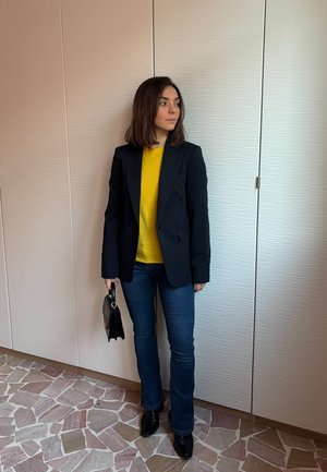 Mujer de pie en el interior, vestida con una blazer negra, suéter amarillo, jeans azules, botas negras, sosteniendo un bolso negro, mirando hacia un lado.