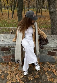 Femeie în pantaloni albi, adidași, palton căprui și șapcă gri stând pe o bancă de piatră într-un parc de toamnă cu frunze împrăștiate, ținând o geantă maro.