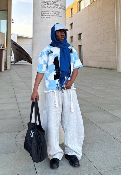 Camisa de cuadros oversized en azul claro y blanco con un motivo gráfico negro, pantalones anchos grises con cordón, bolsa tote negra, bufanda azul, zapatos negros.