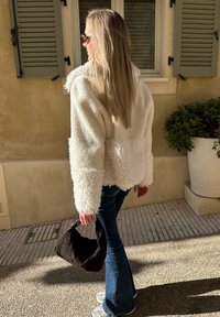 Casaco de fleece branco e fofinho com mangas largas, combinado com jeans azuis flare. A pessoa segura uma bolsa tecida escura, em pé sobre um pavimento texturizado.