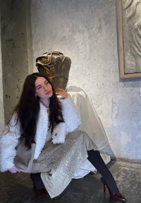 Kvinne sittende i en teksturert gull paljettkjole, hvit pelsjakke i kunstig materiale, og svarte strømpebukser, med burdeos høyhælte sko, med kunst i bakgrunnen.