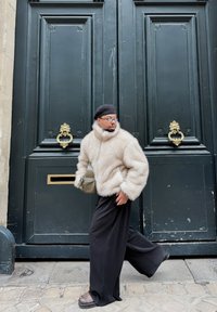 Veste en fausse fourrure beige, pantalon large noir, béret noir et lunettes ; debout près de grandes portes vert foncé avec des ferrures dorées.
