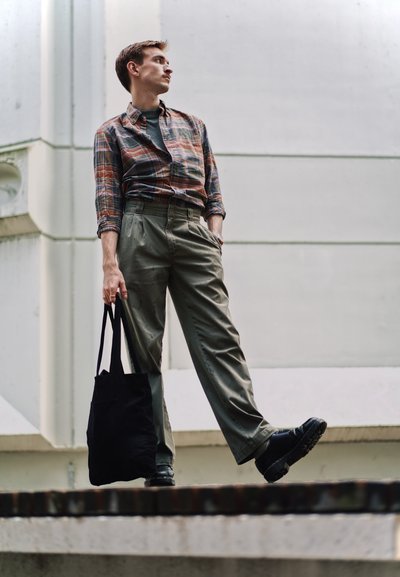 Modelo vistiendo una camisa a cuadros de botones, pantalones de tiro alto verde oliva y zapatos negros, sosteniendo una bolsa de lona negra.