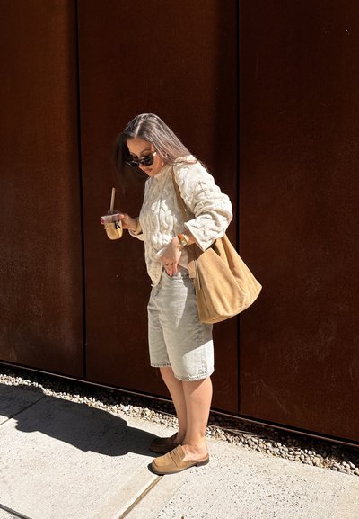 Mujer con gafas de sol, suéter beige y pantalones cortos, sosteniendo café con hielo y un gran bolso marrón claro, de pie en una acera iluminada por el sol junto a una pared de color óxido.