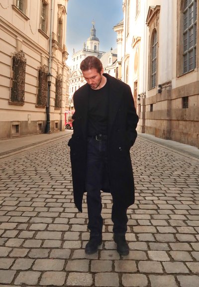 Abrigo negro de doble faz, camiseta oscura, jeans oscuros y botas negras en una calle de adoquines, con edificios históricos y un cielo despejado de fondo.