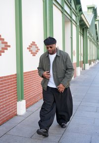 Homme portant un béret noir, des lunettes de soleil, une veste olive, une chemise blanche et un pantalon large noir marchant sur un trottoir pavé à côté d'un bâtiment vert et blanc.