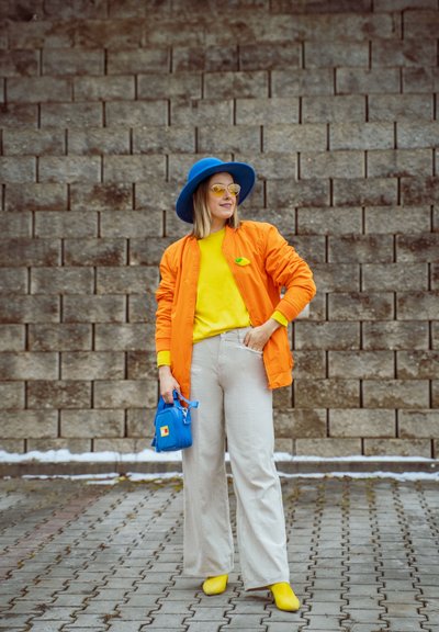 Mujer con sombrero azul brillante, chaqueta naranja, camiseta amarilla y zapatos amarillos sostiene un pequeño bolso azul, de pie en una zona pavimentada con un fondo de pared de ladrillos.