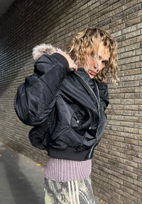 Sort pilotjakke med pelsforet hætte, rynkede ærmer, sølvlynlås og ribbet kant, båret over en stribet sweater og mønstrede bukser.