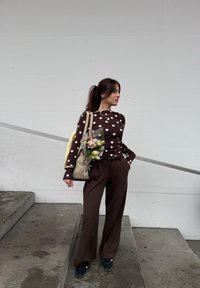 Femme aux cheveux foncés attachés en queue de cheval, portant un haut marron à pois et un pantalon large marron, tenant un sac beige avec des fleurs, debout sur des marches en béton.