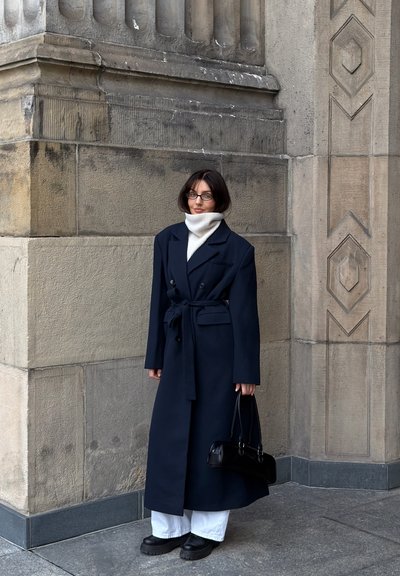 Abrigo azul marino cruzado con cintura ceñida por un cinturón, llevado sobre un jersey de cuello alto blanco, combinado con pantalones blancos y zapatos negros gruesos, sosteniendo un bolso negro.