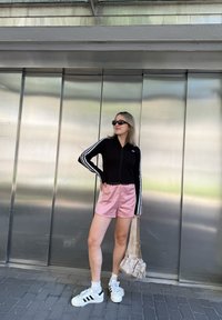 Schwarze Zip-Jacke mit weißen Streifen, pinke Satin-Shorts, weiße Sneakers und eine beige Tasche. Getragen mit Sonnenbrille vor einem metallischen Hintergrund.