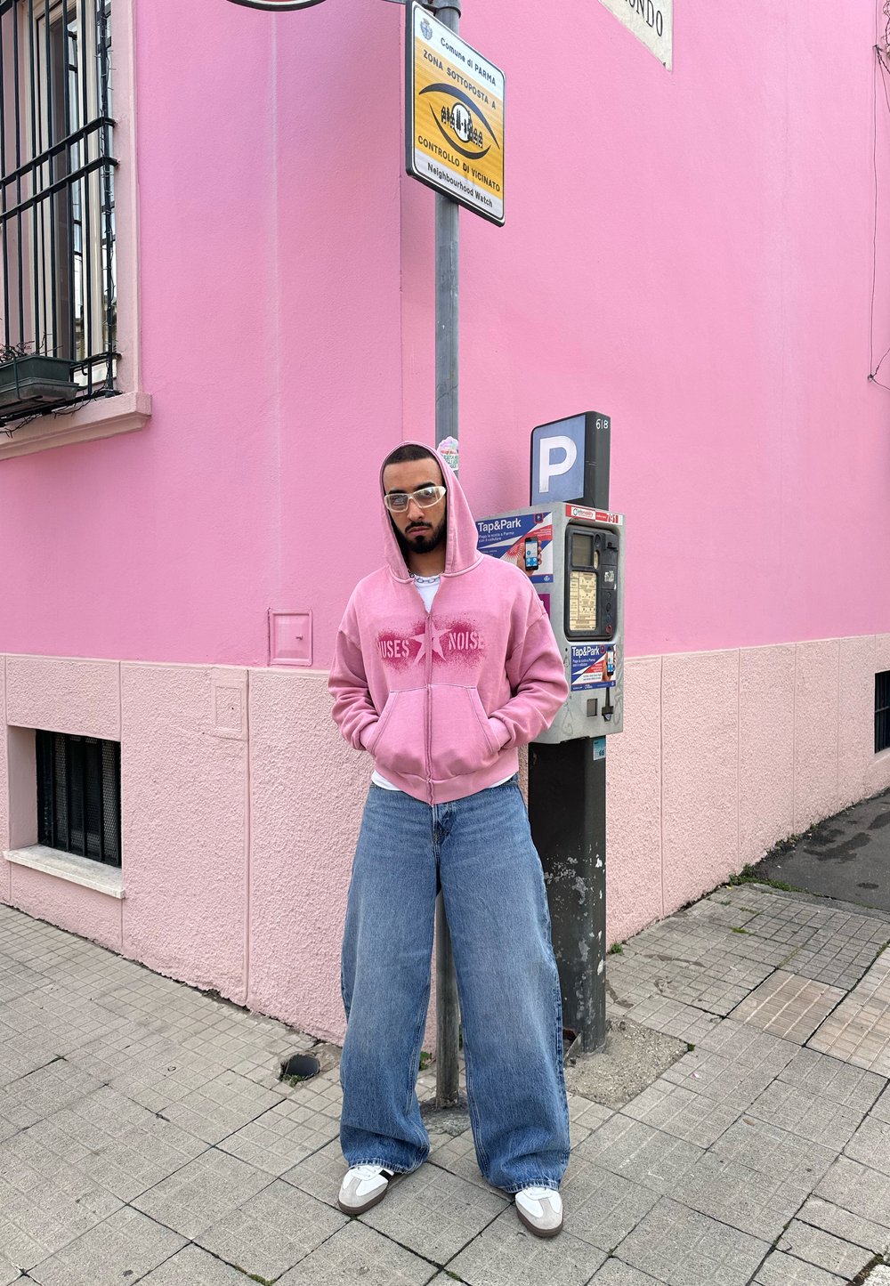 Roza zip-up kapuca s grafiko "NOISE", svetlo modri široki jeans, beli superge in prozorne očala, pred roza zidom.